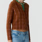 A person wearing the Jane Checker Cardigan by Guest in Residence—a brown, checked lightweight cashmere women’s cardigan—over a green top, paired with blue jeans and a beaded necklace, standing against a plain background.