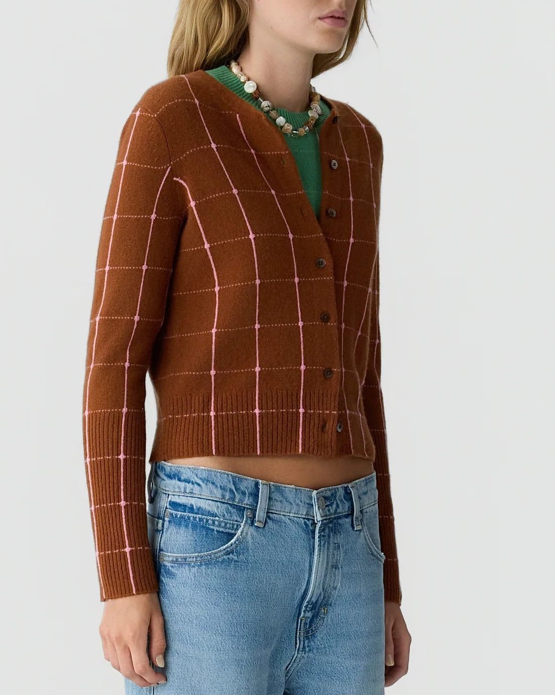 A person wearing the Jane Checker Cardigan by Guest in Residence—a brown, checked lightweight cashmere women’s cardigan—over a green top, paired with blue jeans and a beaded necklace, standing against a plain background.