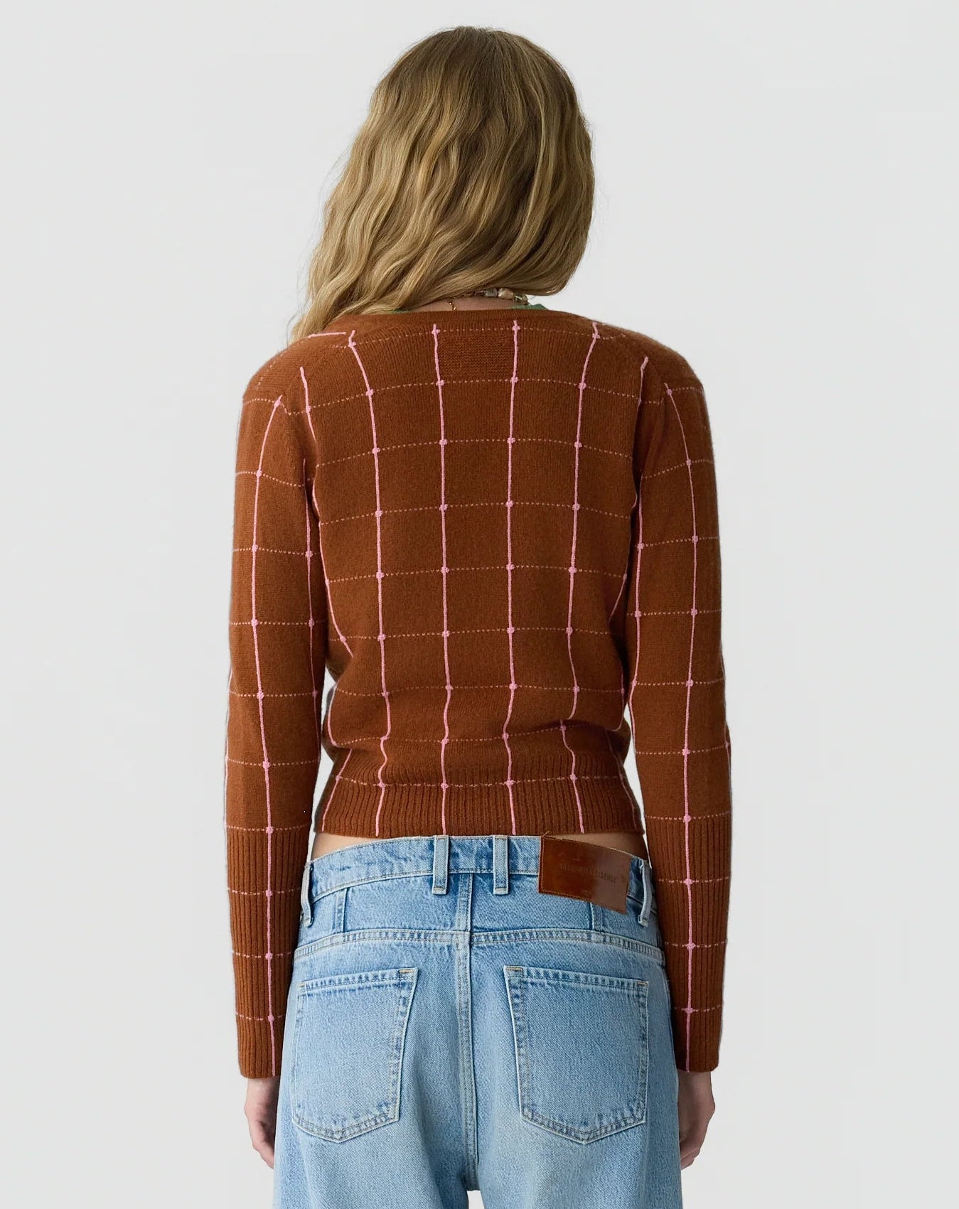 A person with long wavy blonde hair is shown from the back against a plain light background, wearing the Jane Checker Cardigan by Guest in Residence in brown cashmere, paired with light blue jeans.