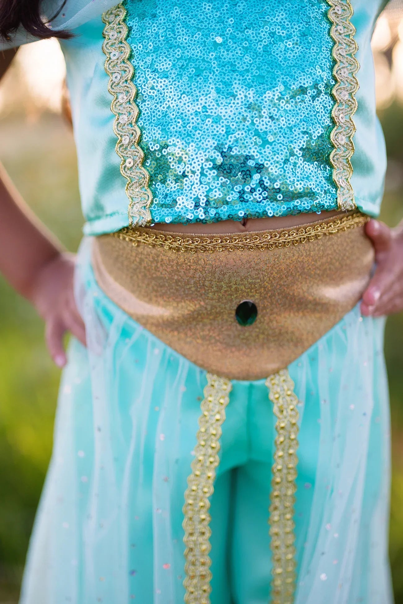 A child dressed in the Great Pretenders Jasmine Princess Set, a premium blue and gold sequined pant costume, stands outdoors with hands on hips; the photo highlights the torso and waist area.