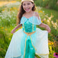 A young child, barefoot and smiling, stands atop a tree stump outdoors surrounded by wildflowers and greenery, holding out the turquoise and gold skirt of the Great Pretenders Jasmine Princess Set—an enchanting costume inspired by a classic pant set—accented with the sparkling charm of emerald hues.