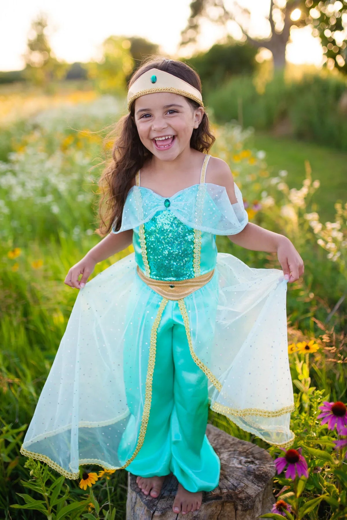 A young child, barefoot and smiling, stands atop a tree stump outdoors surrounded by wildflowers and greenery, holding out the turquoise and gold skirt of the Great Pretenders Jasmine Princess Set—an enchanting costume inspired by a classic pant set—accented with the sparkling charm of emerald hues.