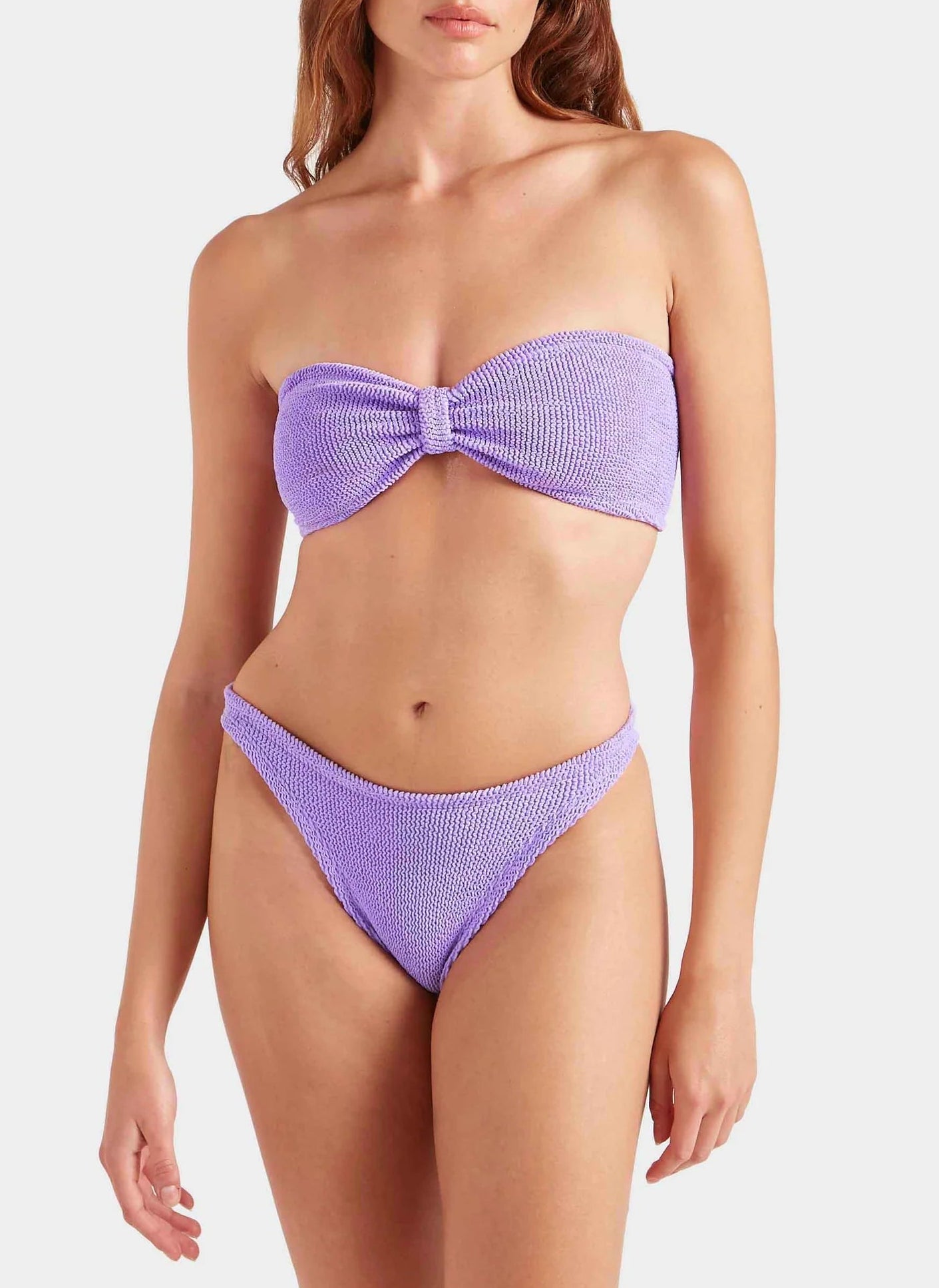 A woman wearing the Hunza G Jean Bikini—a textured, crinkle-fabric bandeau top in lilac with matching high-cut bottoms—stands against a plain white background.
