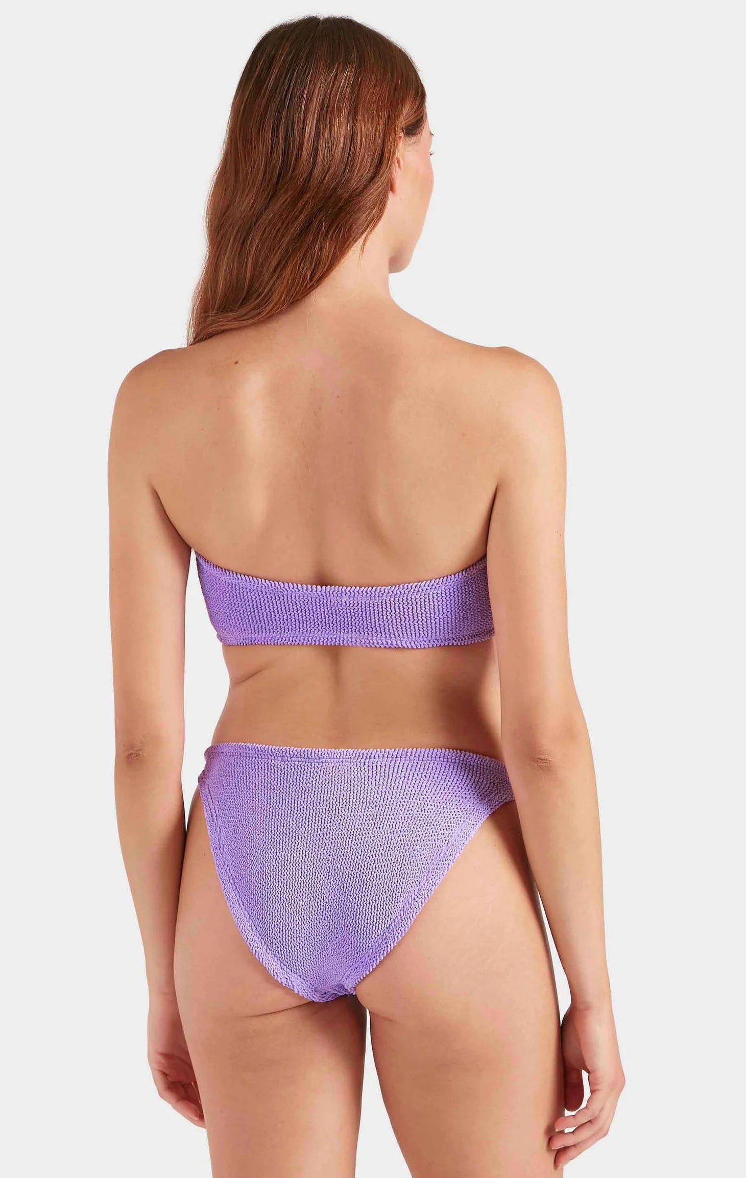 A woman with long brown hair is shown from behind wearing the Jean Bikini by Hunza G, a textured lavender-colored crinkle fabric two-piece, against a white background.