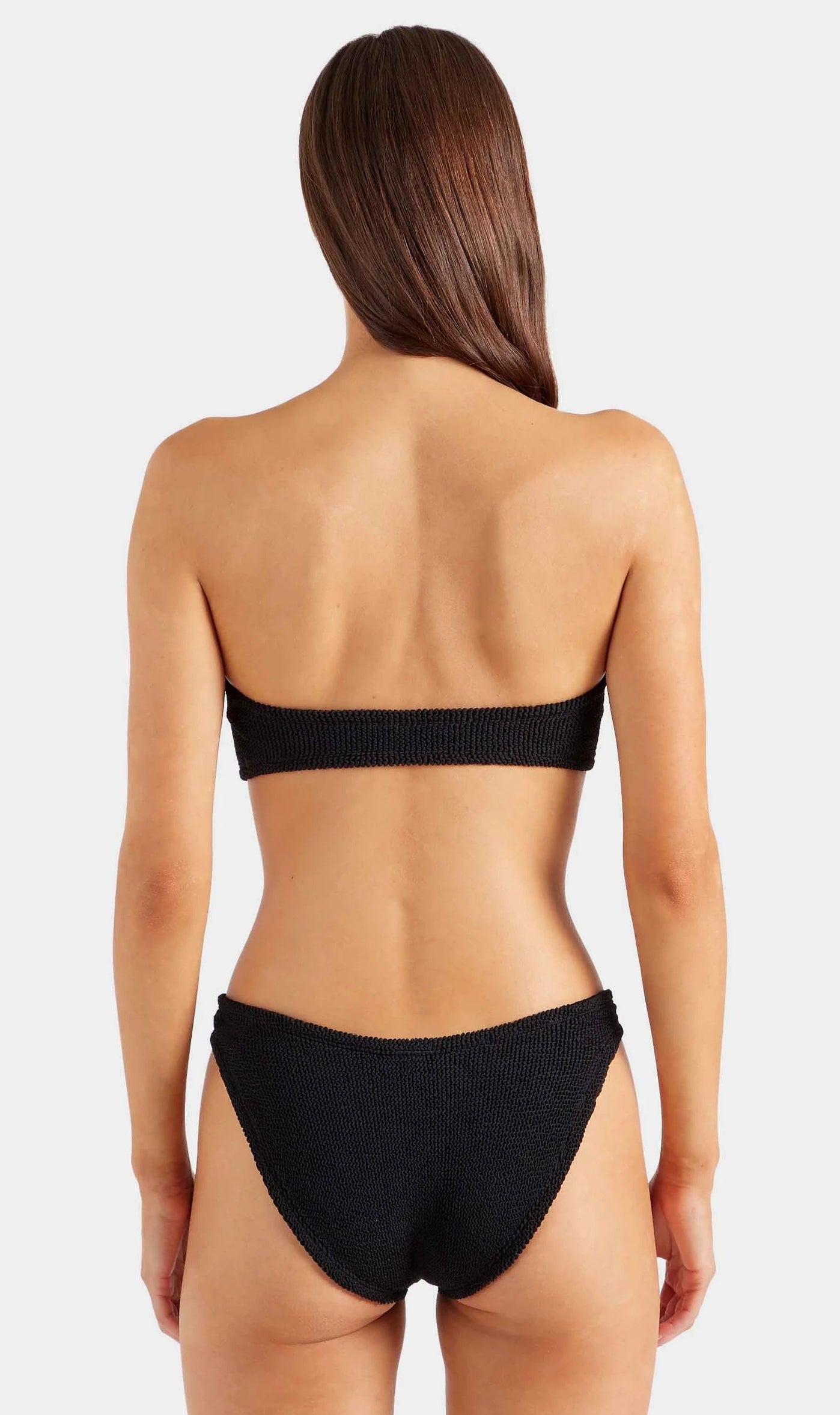 A woman stands with her back to the camera, wearing the Hunza G Jean Bikini—a black crinkle fabric bandeau bikini top with matching bottoms—against a plain white background.