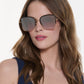 A woman with long brown hair wears Krewe's Jolene Sunglasses, featuring oversized square lenses, and a navy sleeveless top while posing against a plain white background.
