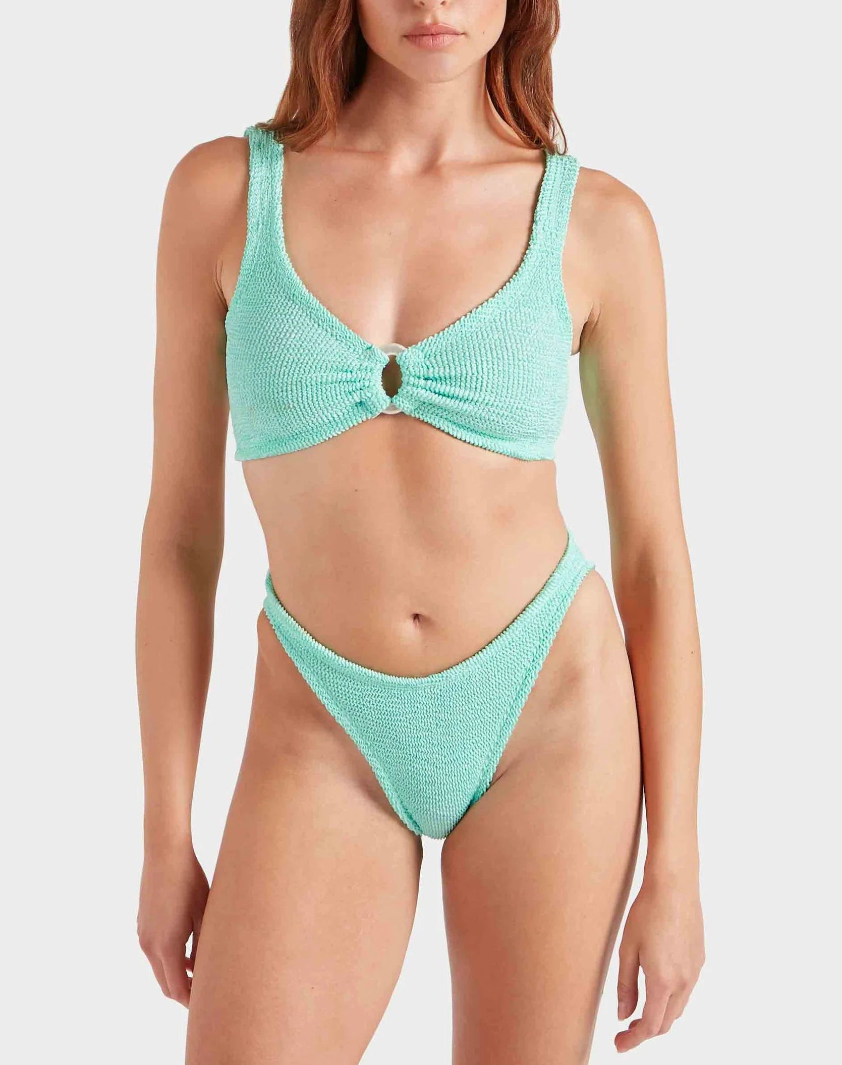 A woman wearing the Hunza G Julia Bikini, featuring a textured mint green, ocean-inspired design with pearly hoop detail on the top and high-cut bottoms, stands against a plain background.