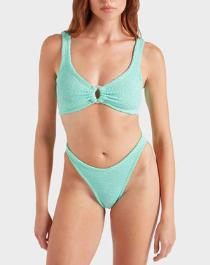 A woman wearing the Hunza G Julia Bikini, featuring a textured mint green, ocean-inspired design with pearly hoop detail on the top and high-cut bottoms, stands against a plain background.