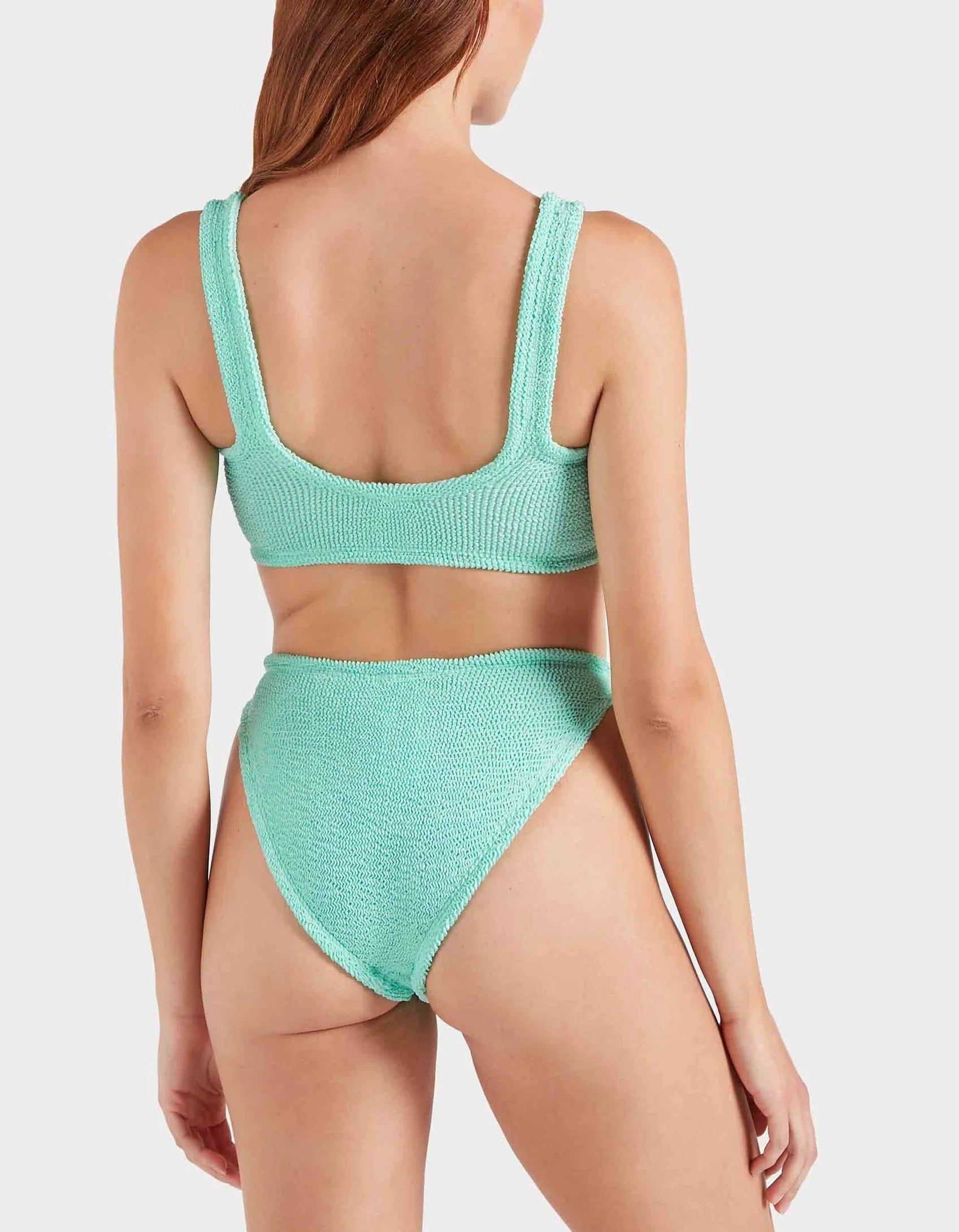 A woman with long hair models the Julia Bikini by Hunza G, featuring an ocean-inspired light blue textured design, shown from the back against a plain light background.