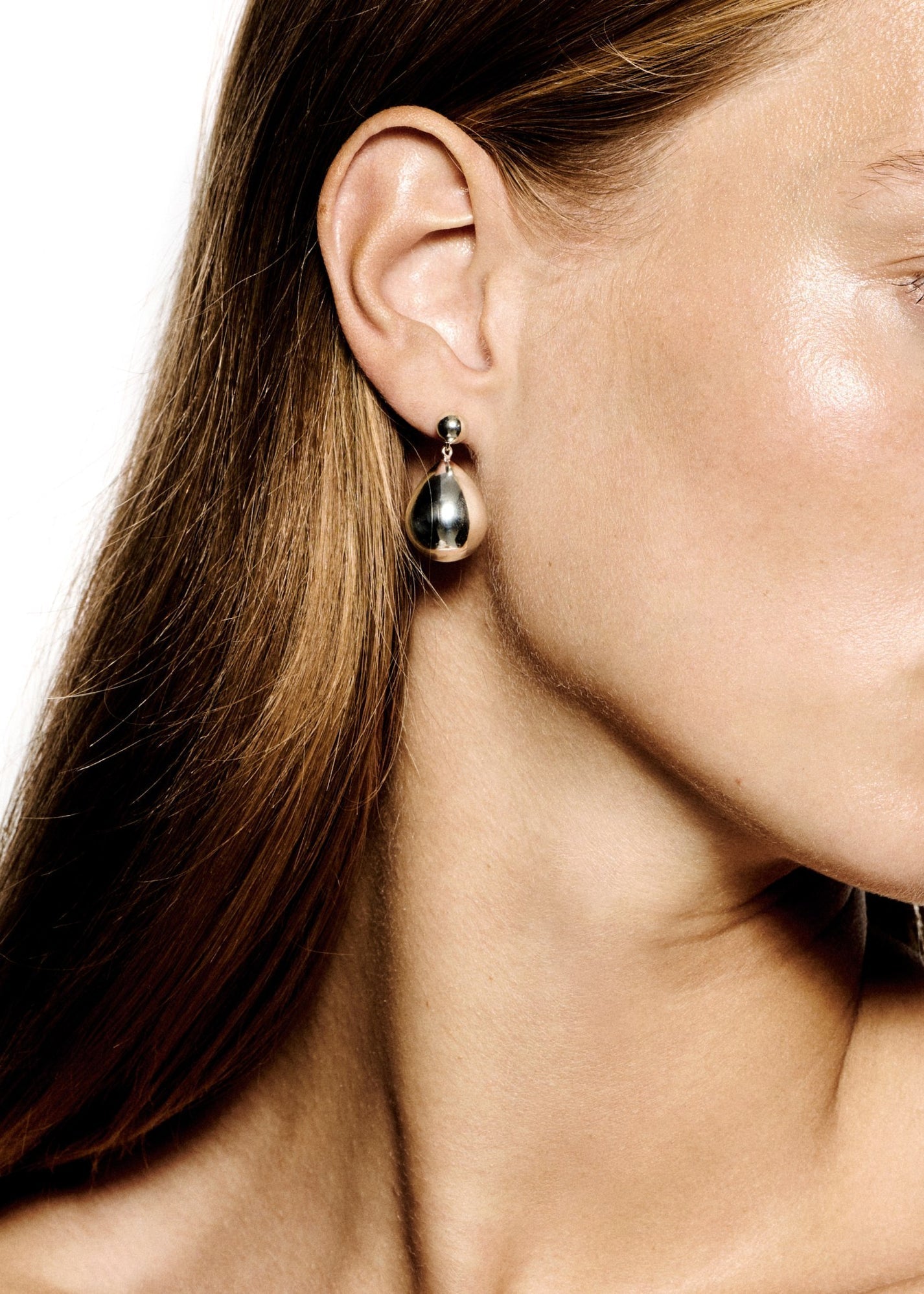 Close-up of a person with straight brown hair wearing the LIÉ STUDIO Julie Earrings from Lie Studio, which are shiny and teardrop-shaped with pearl drops. The skin is smooth and the background is white.