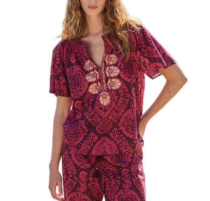 A woman wears the Figue Elena Top, a short-sleeve red and black patterned cotton voile piece with a deep V-neck, paired with matching pants against a white background.