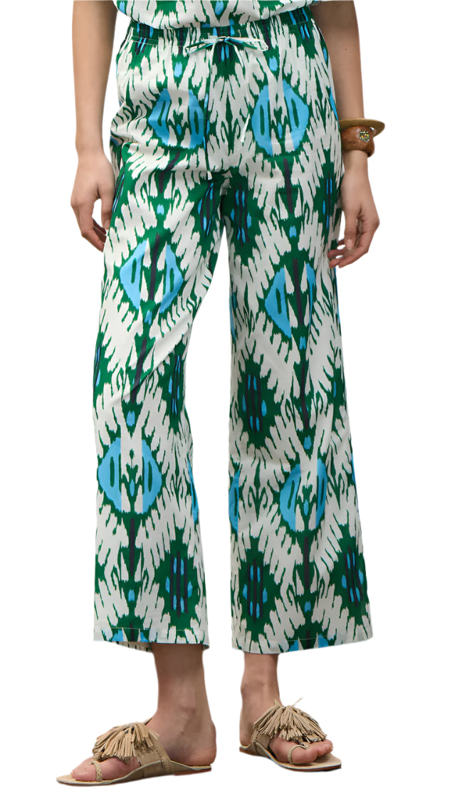 A person stands against a white background wearing the Figue Justine Pant—white with green and blue abstract patterns, side pockets, and a drawstring waist—paired with tan fringe sandals and a brown bracelet.