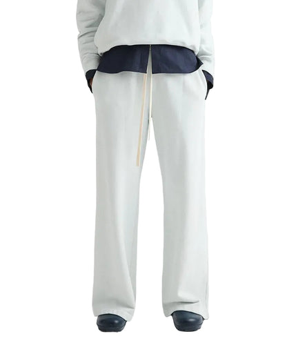 A person wearing TWP PJ Pant—loose-fitting, light gray French terry wide-leg pants with a drawstring—paired with a dark shirt, light top, and dark shoes stands against a white background.