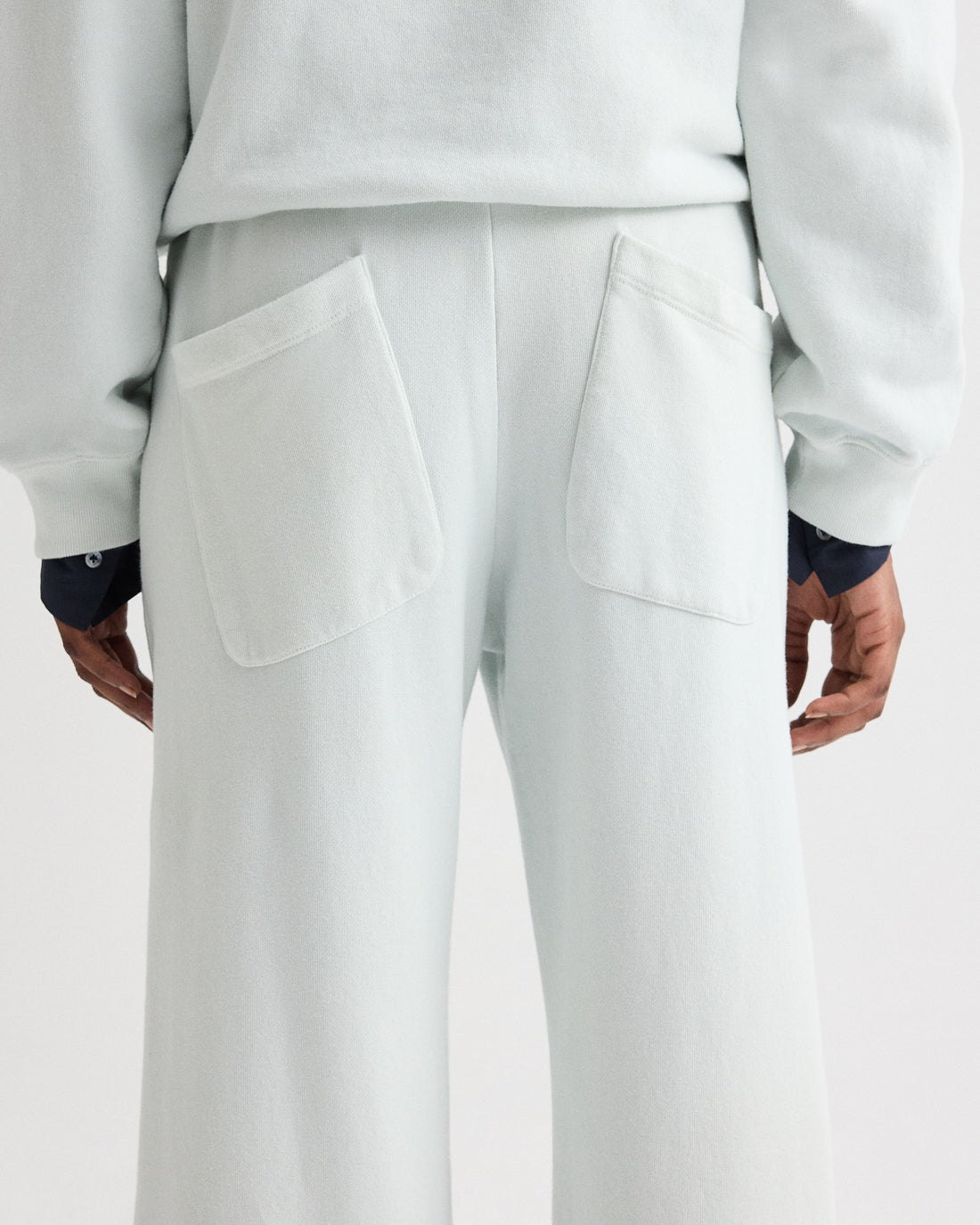 A person is shown from behind wearing TWP's PJ Pant—light-colored, wide-leg pants with two large back pockets, paired with part of a matching top and visible dark shirt cuffs.