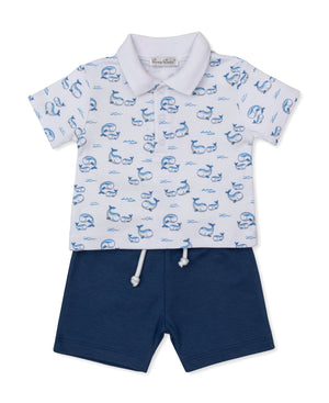 The Kissy Kissy Baby Boys' Whale Pods Bermuda Set, featuring a short-sleeve white polo shirt with a playful whale pods print and dark blue bermuda shorts, is crafted from soft Pima cotton and laid flat on a white background.