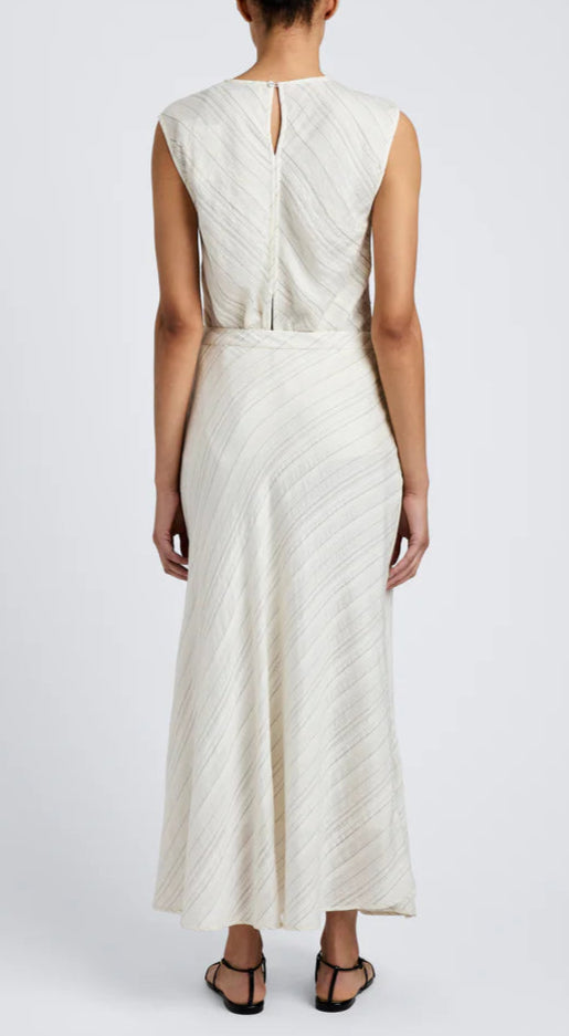 A person stands facing away, wearing the Kaia Woven Stripe Skirt by Proenza Schouler White Label—an off-white, ankle-length skirt with subtle diagonal stripes—paired with black sandals.