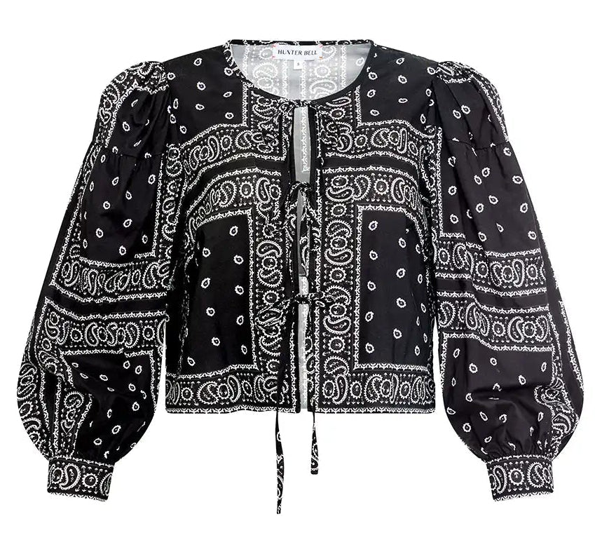 The Katie Top by Hunter Bell NYC is a black bandana-style top with white paisley and geometric prints, balloon sleeves, a round neckline, and a stylish tie-front closure.