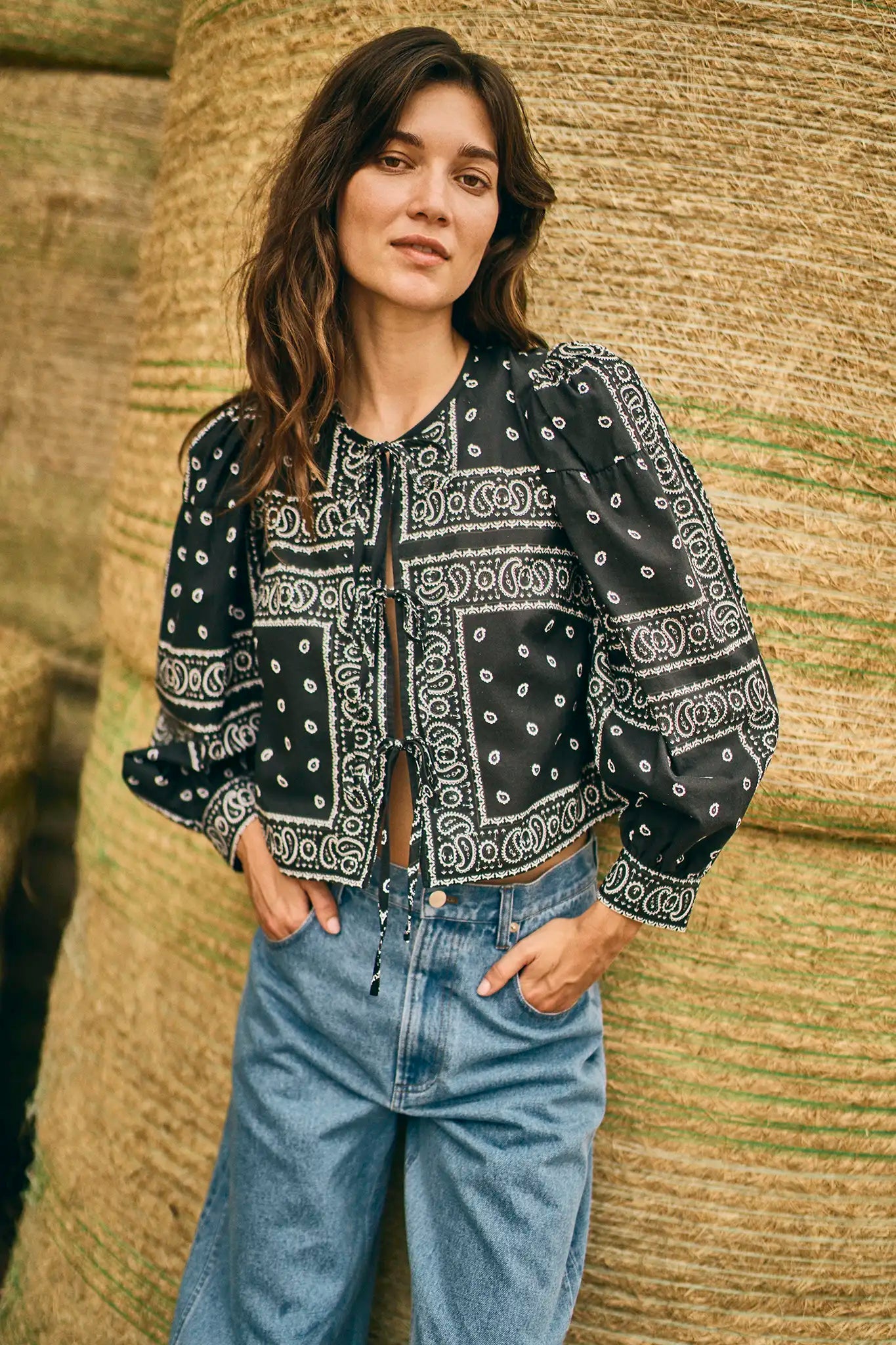 Woman in front of stacked hay bales wears the Hunter Bell NYC "Katie Top," a black bandana-style blouse with balloon sleeves, paired with light blue high-waisted jeans, hands in pockets.