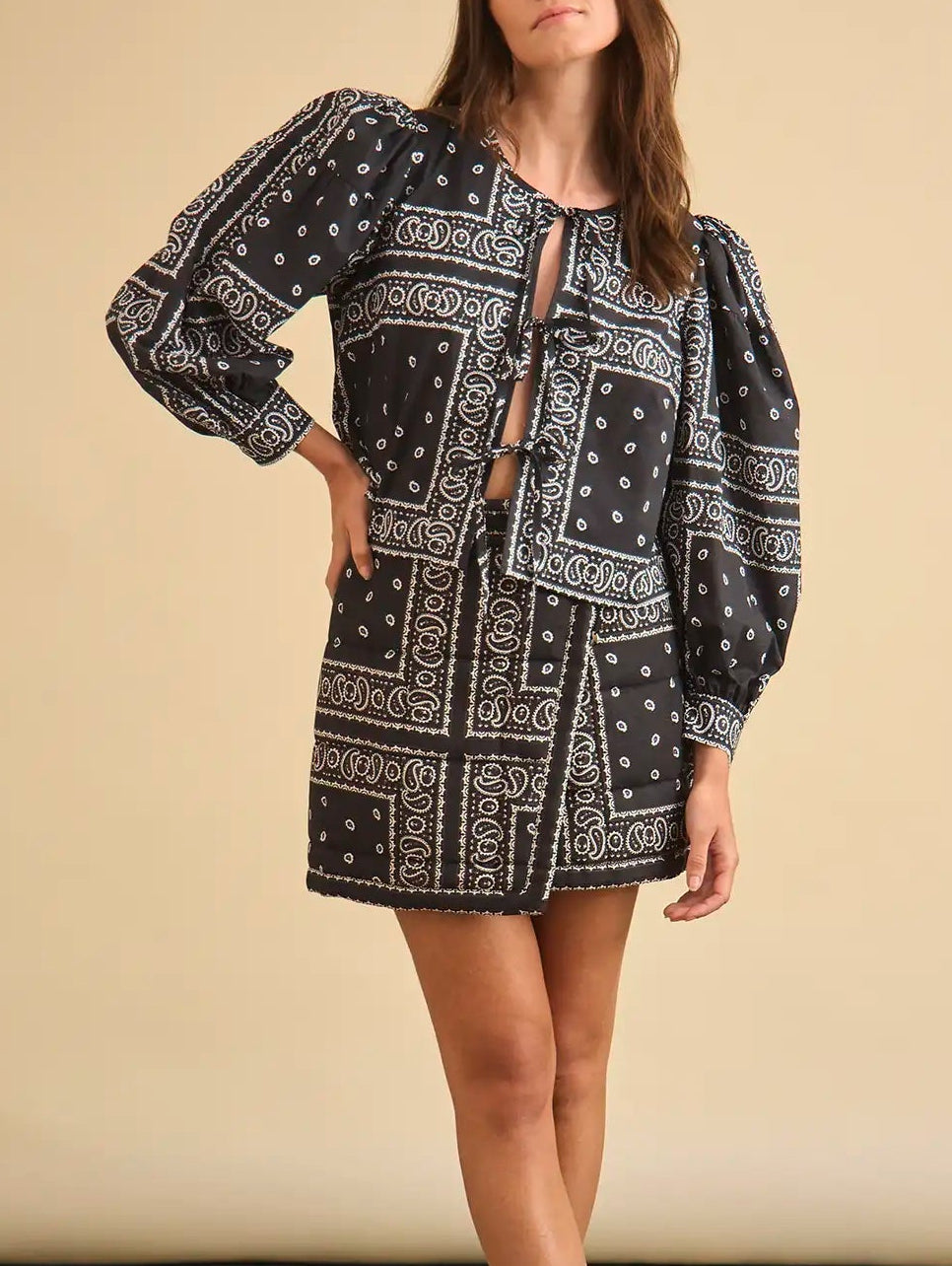 A woman stands against a beige background wearing the Hunter Bell NYC Katie Top, a black bandana top featuring balloon sleeves and a tie-front closure, paired with a matching mini skirt.