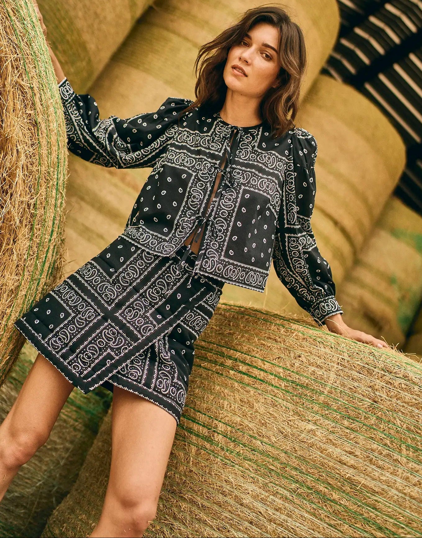 A woman in the black Powell Skirt by Hunter Bell NYC leans against large hay bales in a barn, her statement outfit standing out against the rustic setting.