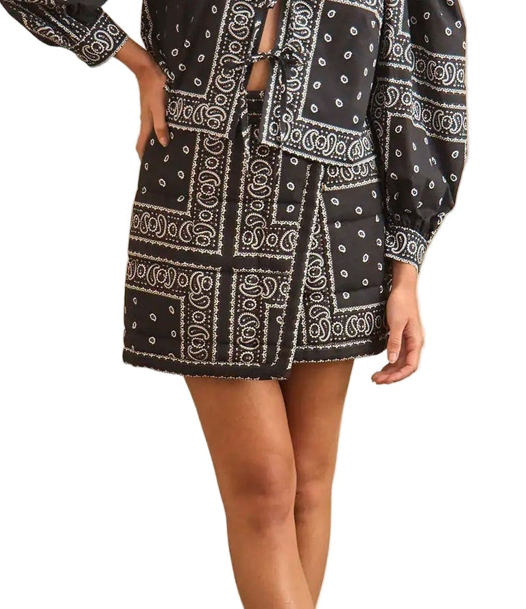 A person wearing the Powell Skirt by Hunter Bell NYC—a black long-sleeve dress with a wrap skirt, front cutout, and a bold Aces Wild bandana-print statement piece tied at the waist.