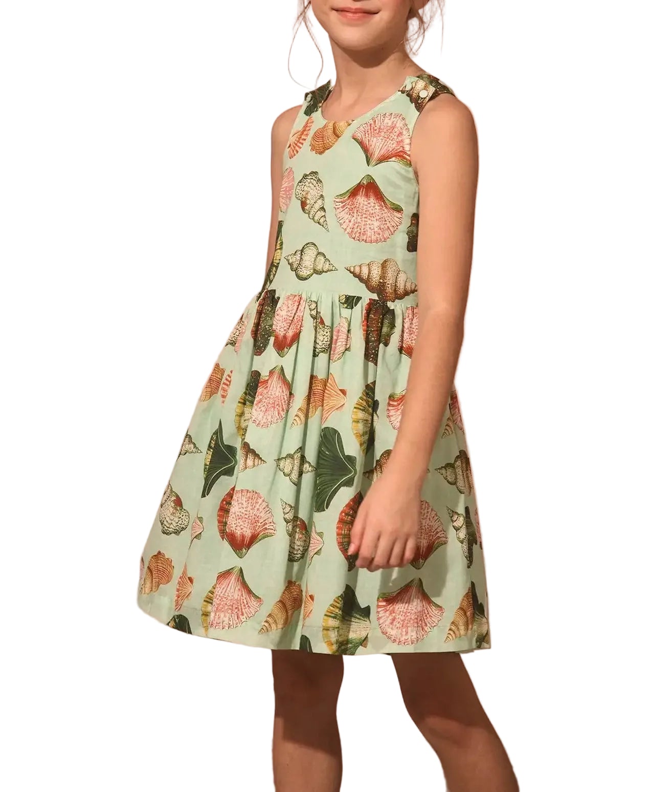 A young girl wears the Girls' June Dress by Hunter Bell NYC, featuring a sleeveless design with a seashell print on a light green background, standing against a white backdrop.