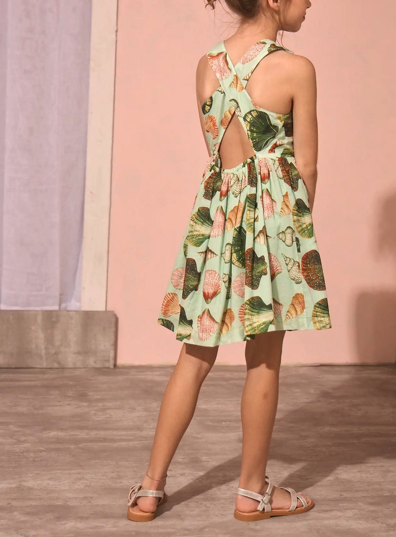 A young girl stands indoors, facing away, wearing the Girls' June Dress by Hunter Bell NYC in the delightful Somers Shell Mint print with crisscross straps, paired with light sandals—a perfect mommy and me summer style.