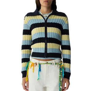 A person wears the Kirby Jacket by Guest in Residence—featuring blue, yellow, and black stripes—paired with white pants and a belt made from colorful braided strings.