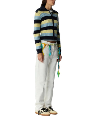 A person wears the Guest in Residence Kirby Jacket—a cropped women's knit zip-up with stripes—paired with white pants, a colorful belt, and beige sneakers with black stripes, all set against a white background.