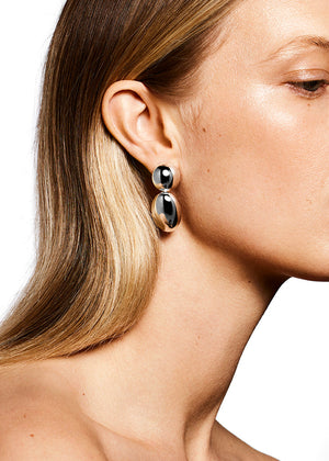 Close-up of a person with long, straight blonde hair wearing shiny, oval-shaped LIÉ STUDIO Klara Earrings made from 925 sterling silver. The background is plain white.