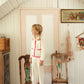 A child dressed in white wears Louise Misha’s Girls’ Salia Pants, standing on a wooden chair in a vintage room with white paneled walls, floral decor, and another empty chair nearby.