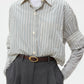 A person wears the TWP Next Ex Dancing Striped Cotton Shirt over a gray long-sleeve top, tucked into high-waisted gray trousers with a brown belt for a boxy silhouette.