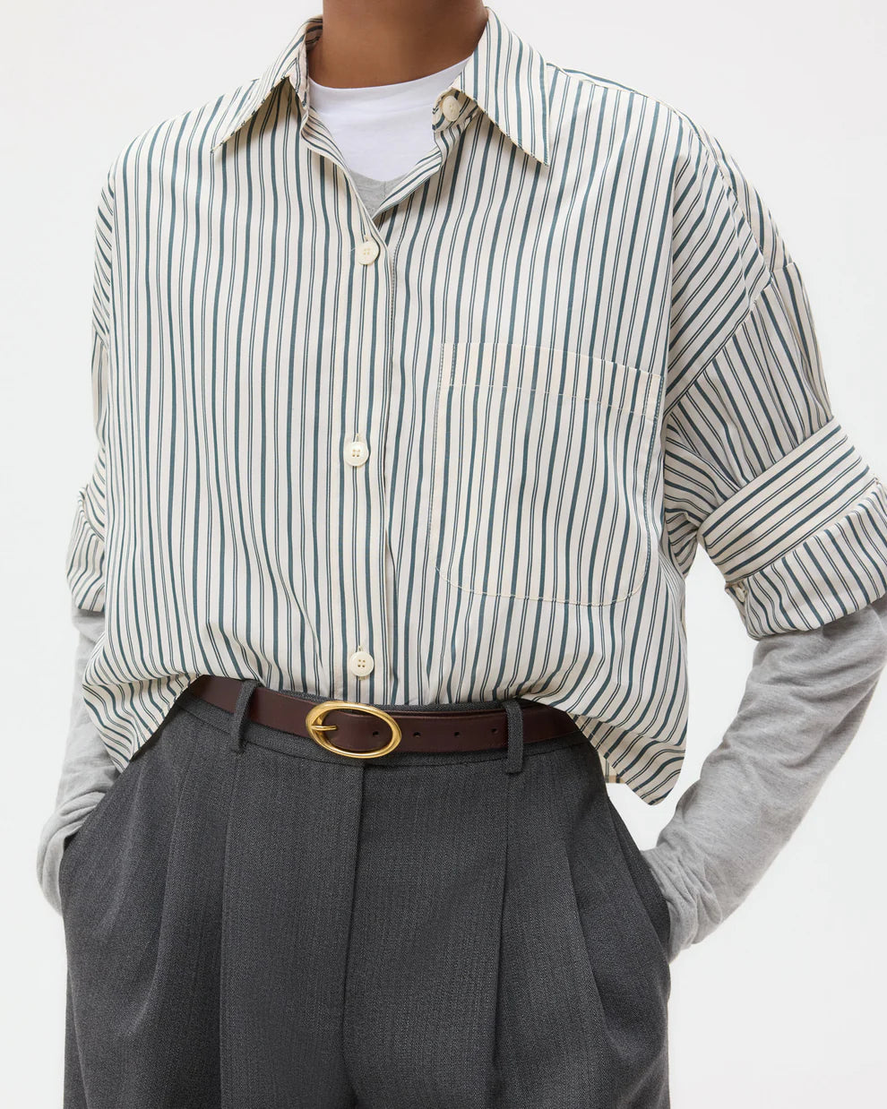 A person wears the TWP Next Ex Dancing Striped Cotton Shirt over a gray long-sleeve top, tucked into high-waisted gray trousers with a brown belt for a boxy silhouette.