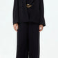 A person wears the TWP Boy Blazer, a loose-fitting black wool crepe suit with wide-leg trousers, a matching black top, and a gold belt, standing against a plain light background.