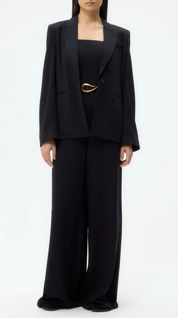 A person wears the TWP Boy Blazer, a loose-fitting black wool crepe suit with wide-leg trousers, a matching black top, and a gold belt, standing against a plain light background.