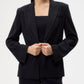 A person wears the TWP Boy Blazer and matching black pants, holding the blazer’s front button with both hands. The face is partially out of frame.