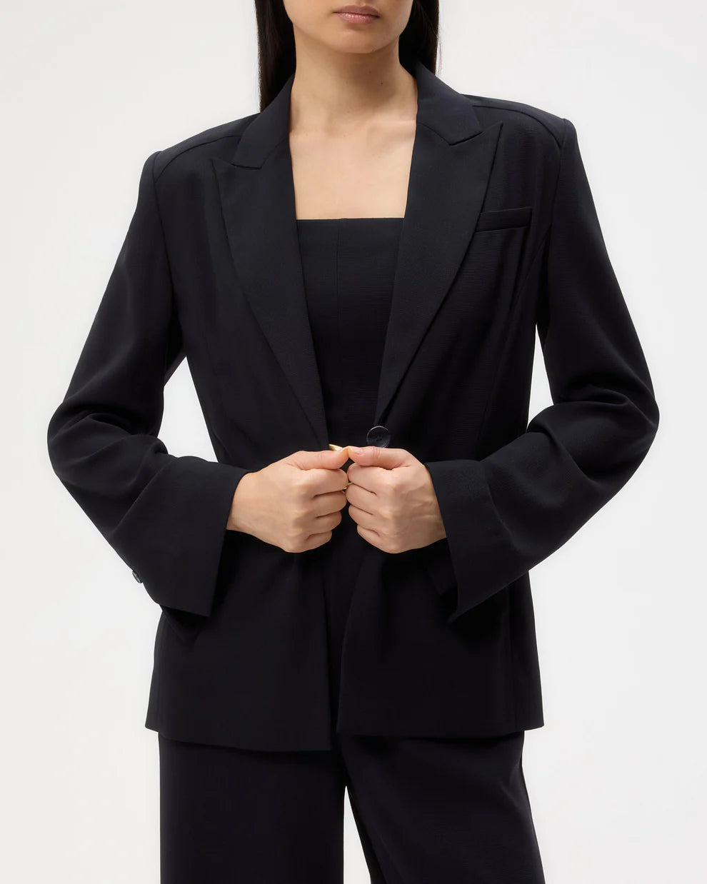 A person wears the TWP Boy Blazer and matching black pants, holding the blazer’s front button with both hands. The face is partially out of frame.