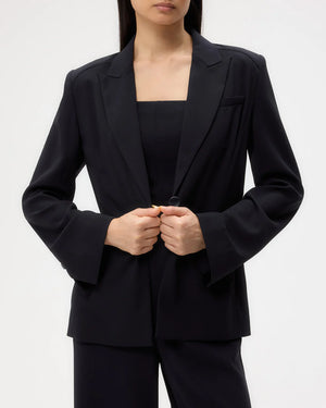A person wears the TWP Boy Blazer and matching black pants, holding the blazer’s front button with both hands. The face is partially out of frame.