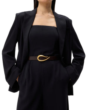 A woman wears the TWP Boy Blazer over a strapless top, styled with a black belt featuring a large gold abstract buckle, hands in pockets, against a plain background.
