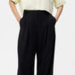 A person wearing a light yellow button-up shirt, oversized TWP Carries Trouser in black wool crepe, and brown shoes stands against a plain white background.