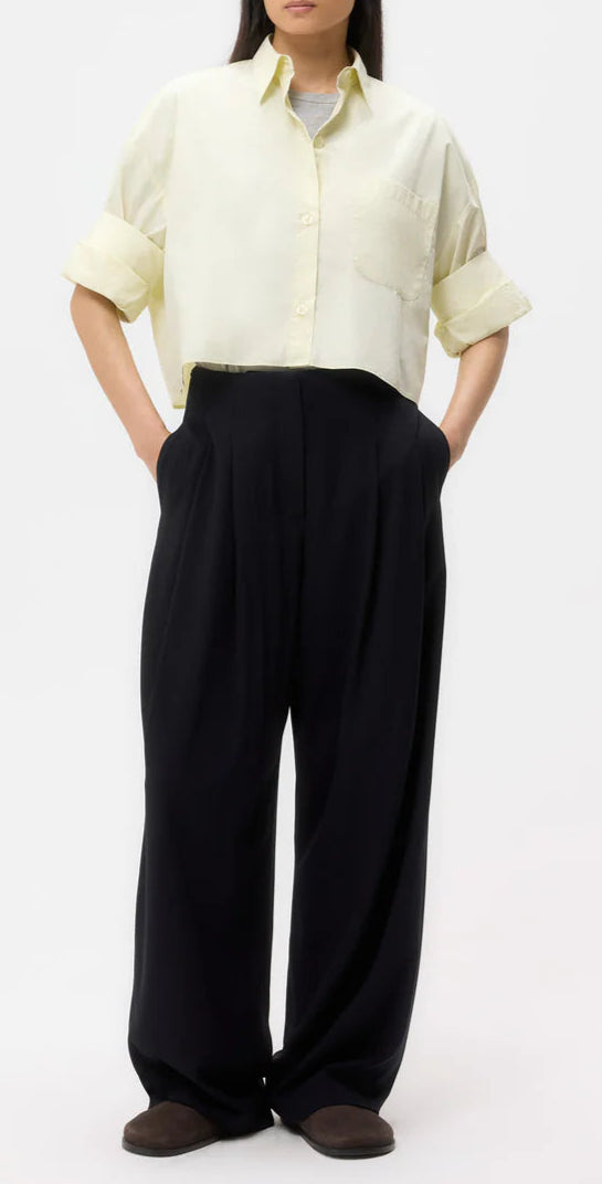 A person wearing a light yellow button-up shirt, oversized TWP Carries Trouser in black wool crepe, and brown shoes stands against a plain white background.