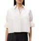 A person stands against a plain background wearing the TWP Next Ex Shirt, a vintage-washed poplin cropped button-down with cuffed sleeves and a large front pocket, paired with dark high-waisted pants.