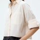 A woman wears the TWP Next Ex Shirt in light beige, featuring a cropped, boxy fit with rolled sleeves and button-down front, paired with black high-waisted trousers against a plain background.
