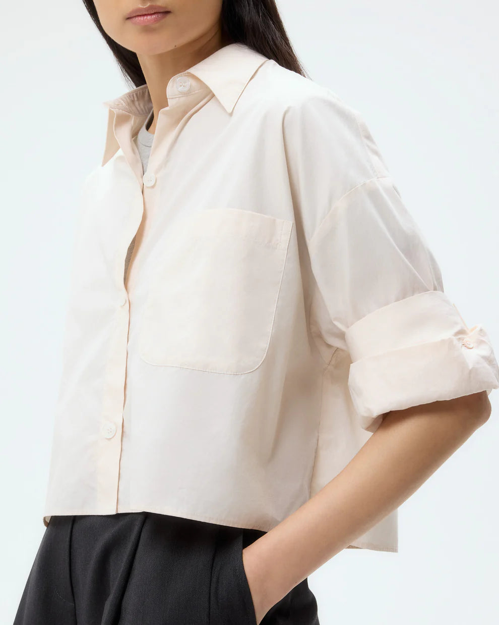 A woman wears the TWP Next Ex Shirt in light beige, featuring a cropped, boxy fit with rolled sleeves and button-down front, paired with black high-waisted trousers against a plain background.