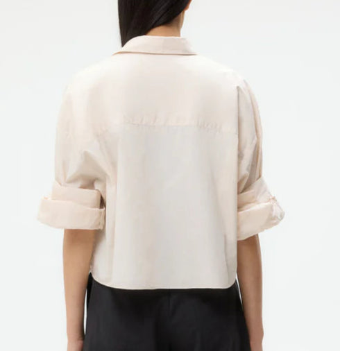 A person is shown from the back wearing the TWP Next Ex Shirt, a light beige, short-sleeved collared top with a boxy silhouette and rolled sleeves for a modern look, styled with black pants.