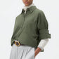 A person stands against a plain background wearing TWP’s Next Ex Shirt in vintage-washed green over a white top, paired with wide-leg light gray trousers for a boxy silhouette and tan shoes.