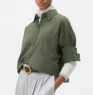 A person stands against a plain background wearing TWP’s Next Ex Shirt in vintage-washed green over a white top, paired with wide-leg light gray trousers for a boxy silhouette and tan shoes.