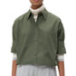 A person in the TWP Next Ex Shirt, a vintage washed poplin green boxy button-up, layered over a white turtleneck and light grey high-waisted trousers, stands against a plain white background.