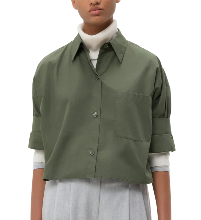 A person in the TWP Next Ex Shirt, a vintage washed poplin green boxy button-up, layered over a white turtleneck and light grey high-waisted trousers, stands against a plain white background.