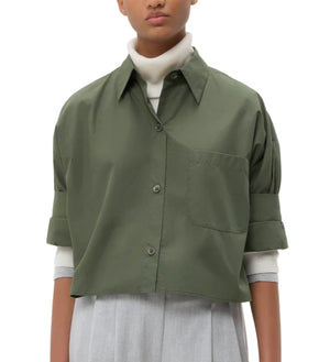 A person in the TWP Next Ex Shirt, a vintage washed poplin green boxy button-up, layered over a white turtleneck and light grey high-waisted trousers, stands against a plain white background.